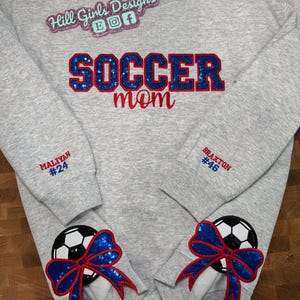 May include: A light gray crewneck sweatshirt featuring the text "SOCCER mom" in blue sequin and red embroidery. The sleeves are personalized with "MALIYAH #24" and "BRAXTON #46" in red and blue. Two soccer ball patches with blue sequin bows are attached.