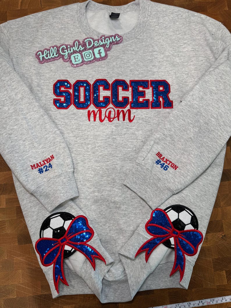 May include: A light gray crewneck sweatshirt featuring the text "SOCCER mom" in blue sequin and red embroidery. The sleeves are personalized with "MALIYAH #24" and "BRAXTON #46" in red and blue. Two soccer ball patches with blue sequin bows are attached.