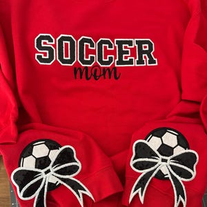 May include: A bright red crewneck sweatshirt featuring the word SOCCER in large black block letters with white outlines, with the word mom in black script below. Each sleeve cuff displays a black and white soccer ball patch with a black bow.