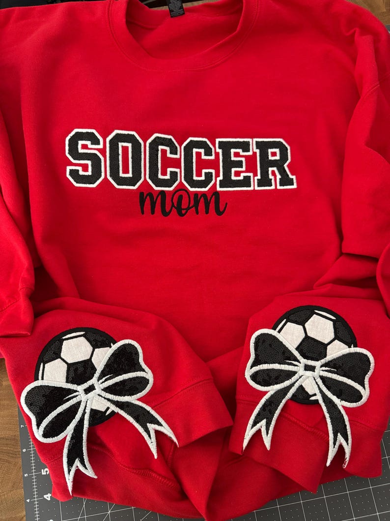 May include: A bright red crewneck sweatshirt featuring the word SOCCER in large black block letters with white outlines, with the word mom in black script below. Each sleeve cuff displays a black and white soccer ball patch with a black bow.