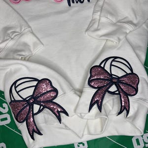 Puede incluir: Una sudadera blanca de manga larga con el texto Volleyball mom bordado en azul oscuro y rosa en el pecho. Cada pu&ntilde;o de la manga muestra un gr&aacute;fico de voleibol bordado en azul oscuro con un detalle de lazo de lentejuelas rosa.