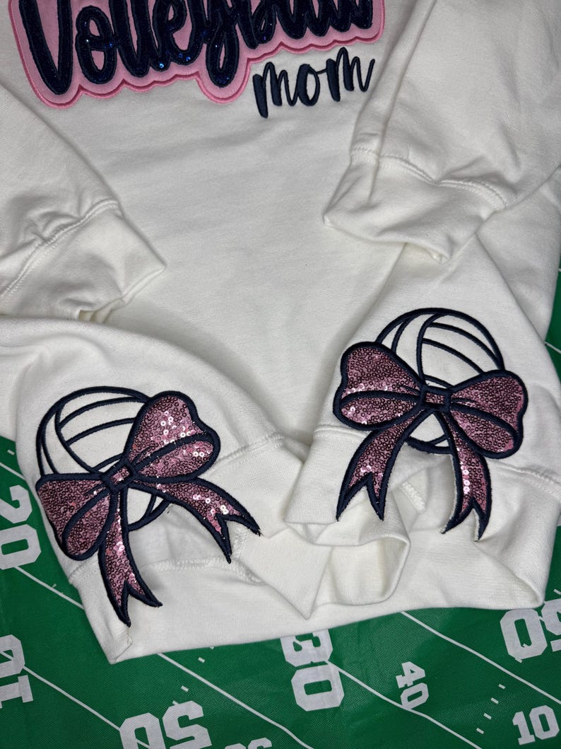 Puede incluir: Una sudadera blanca de manga larga con el texto Volleyball mom bordado en azul oscuro y rosa en el pecho. Cada pu&ntilde;o de la manga muestra un gr&aacute;fico de voleibol bordado en azul oscuro con un detalle de lazo de lentejuelas rosa.