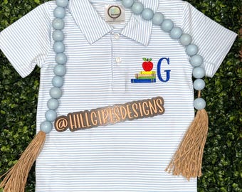 May include: A light blue and white striped polo shirt with an embroidered apple, books, and the letter "G". The shirt has a three-button placket and short sleeves. A light blue beaded garland with tassels is draped across the shirt.