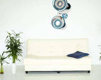 May include: A white couch with a blue pillow in front of a white wall with a blue and white abstract wall art piece.
