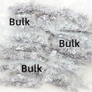 May include: A collection of small, silver metal letter charms in clear plastic bags. The word "Bulk" is printed in black on some of the bags. The charms appear to be individual letters of the alphabet.