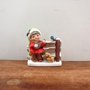 May include: A ceramic figurine of a child wearing a green and white hat playing a banjo with a small dog and a blue bird perched on a fence in a snowy scene.