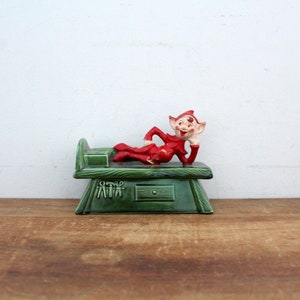 May include: A vintage ceramic elf figurine in a red outfit, reclining on a green dresser. The dresser has a drawer and decorative tools. The elf is smiling, with a hand under its chin. A whimsical holiday decoration.