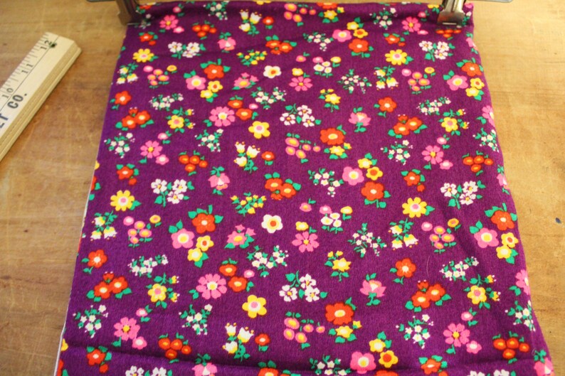 May include: Purple fabric with a small floral pattern in shades of pink, yellow, red, and green.