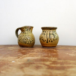 May include: Two small, handcrafted ceramic vases with a rustic aesthetic. The vases are glazed in shades of brown and tan, with dark brown drip and speckled patterns. One has a handle. They sit on a wooden surface.