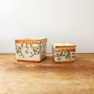 May include: Two square ceramic boxes with floral and ribbon designs. The larger box is white with brown ribbons and colorful flowers. The smaller box is white with brown ribbons and pink flowers.