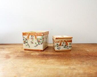 May include: Two square ceramic boxes with floral and ribbon designs. The larger box is white with brown ribbons and colorful flowers. The smaller box is white with brown ribbons and pink flowers.