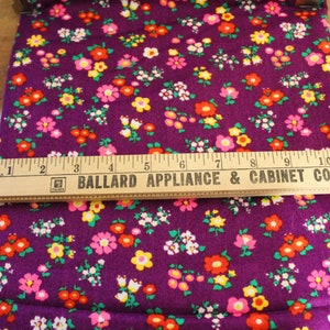 May include: Purple fabric with a small floral print in shades of pink, yellow, red, and green. A wooden ruler with the text "BALLARD APPLIANCE & CABINET CO." is placed on top of the fabric.