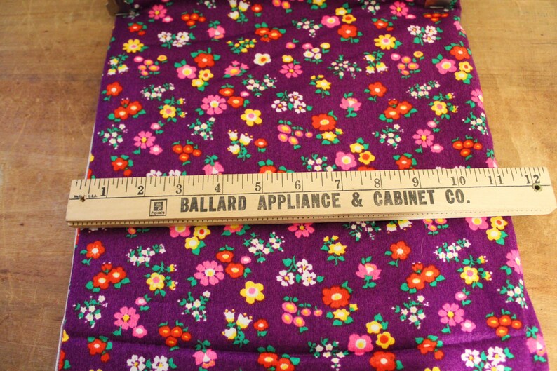 May include: Purple fabric with a small floral print in shades of pink, yellow, red, and green. A wooden ruler with the text "BALLARD APPLIANCE & CABINET CO." is placed on top of the fabric.