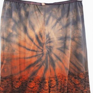 May include: A long, flowing skirt with a tie-dye pattern in shades of orange, brown, and gray. The skirt features a spiral design and a decorative lace trim at the hem. The fabric appears lightweight and suitable for warm weather.