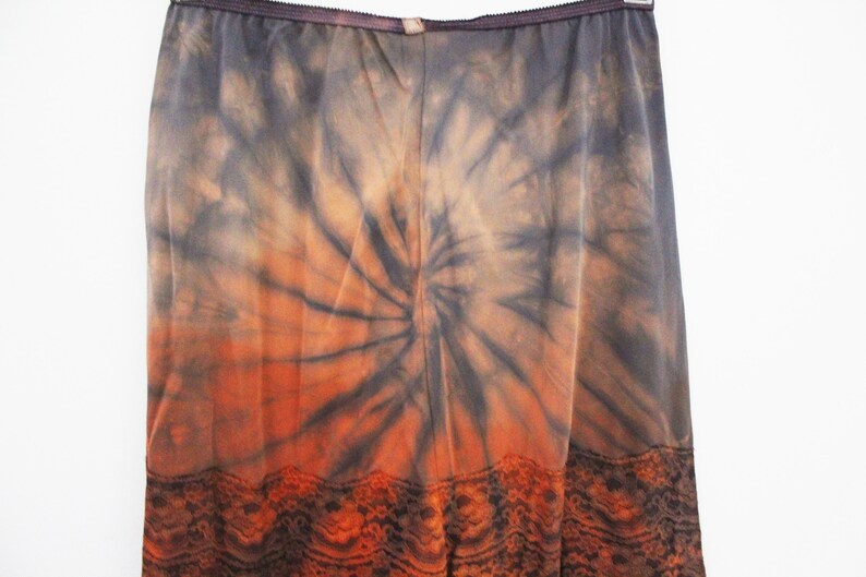 May include: A long, flowing skirt with a tie-dye pattern in shades of orange, brown, and gray. The skirt features a spiral design and a decorative lace trim at the hem. The fabric appears lightweight and suitable for warm weather.