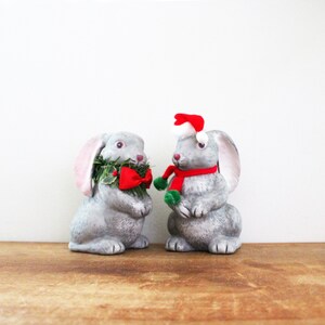 May include: Two gray rabbit figurines. One wears a green wreath and red bow, the other a red scarf and Santa hat. The bunnies are festive holiday decorations, perfect for Christmas or Easter.