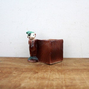 May include: A ceramic planter shaped like a small boy with a green hat, holding a brown rectangular planter.