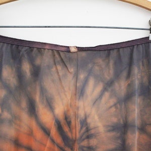 May include: A pair of tie-dye pants in shades of gray and orange, hanging on a wooden hanger. The pants have a dark waistband and a unique, swirling pattern.
