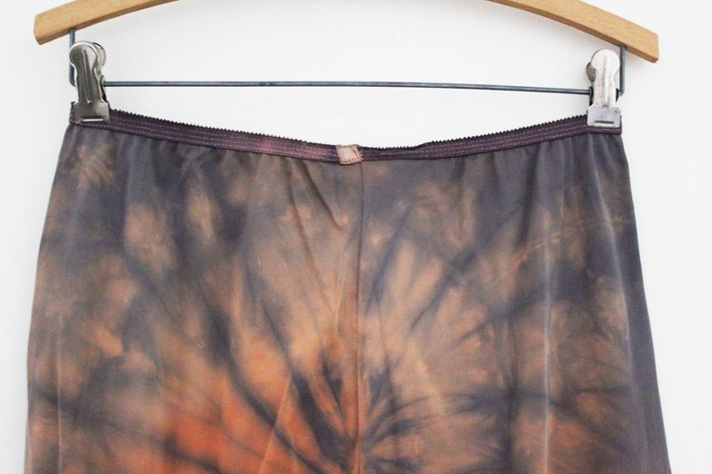 May include: A pair of tie-dye pants in shades of gray and orange, hanging on a wooden hanger. The pants have a dark waistband and a unique, swirling pattern.
