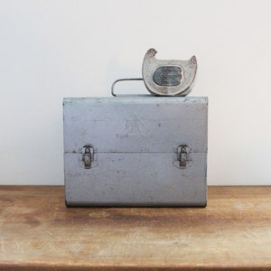 May include: A vintage metal lunchbox with a gray finish and two latches. The lunchbox has a small, gray, ceramic chicken figurine on top.