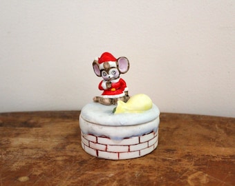 May include: A ceramic Christmas trinket box featuring a small mouse wearing a Santa hat and holding a sack of gifts. The box is decorated with a brick chimney and snow.