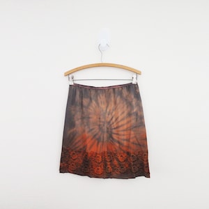 May include: A knee-length skirt with a tie-dye pattern in shades of brown and orange, hanging on a wooden hanger. The skirt has a dark brown lace trim at the bottom and a brown waistband.