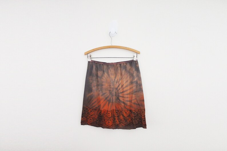 May include: A knee-length skirt with a tie-dye pattern in shades of brown and orange, hanging on a wooden hanger. The skirt has a dark brown lace trim at the bottom and a brown waistband.