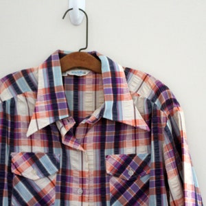 May include: A long-sleeved plaid shirt with a white, orange, purple, and blue plaid pattern. The shirt has a button-down collar and two chest pockets.