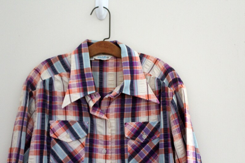 May include: A long-sleeved plaid shirt with a white, orange, purple, and blue plaid pattern. The shirt has a button-down collar and two chest pockets.