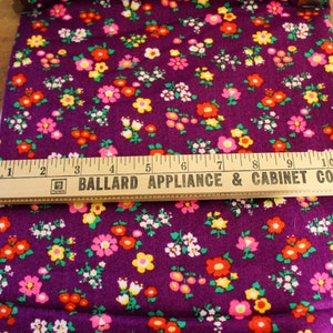 May include: Purple fabric with a repeating pattern of small, colorful flowers. A wooden ruler with the text "Ballard Appliance & Cabinet Co." is placed on top of the fabric.