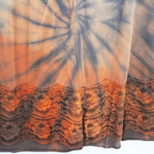 May include: A tie-dye fabric with a spiral pattern in shades of orange, gray, and blue. The bottom edge features a decorative lace trim in shades of brown and orange. The fabric appears to be a dress or skirt.