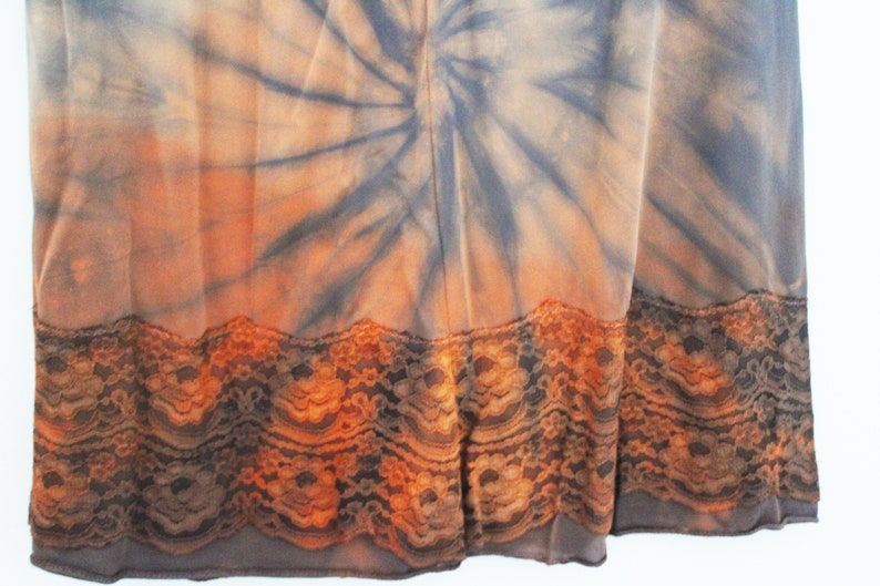 May include: A tie-dye fabric with a spiral pattern in shades of orange, gray, and blue. The bottom edge features a decorative lace trim in shades of brown and orange. The fabric appears to be a dress or skirt.