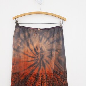 May include: A tie-dye skirt in shades of orange, brown, and gray, hanging on a wooden hanger. The skirt features a spiral pattern and a lace trim at the hem. The garment is displayed against a white background.