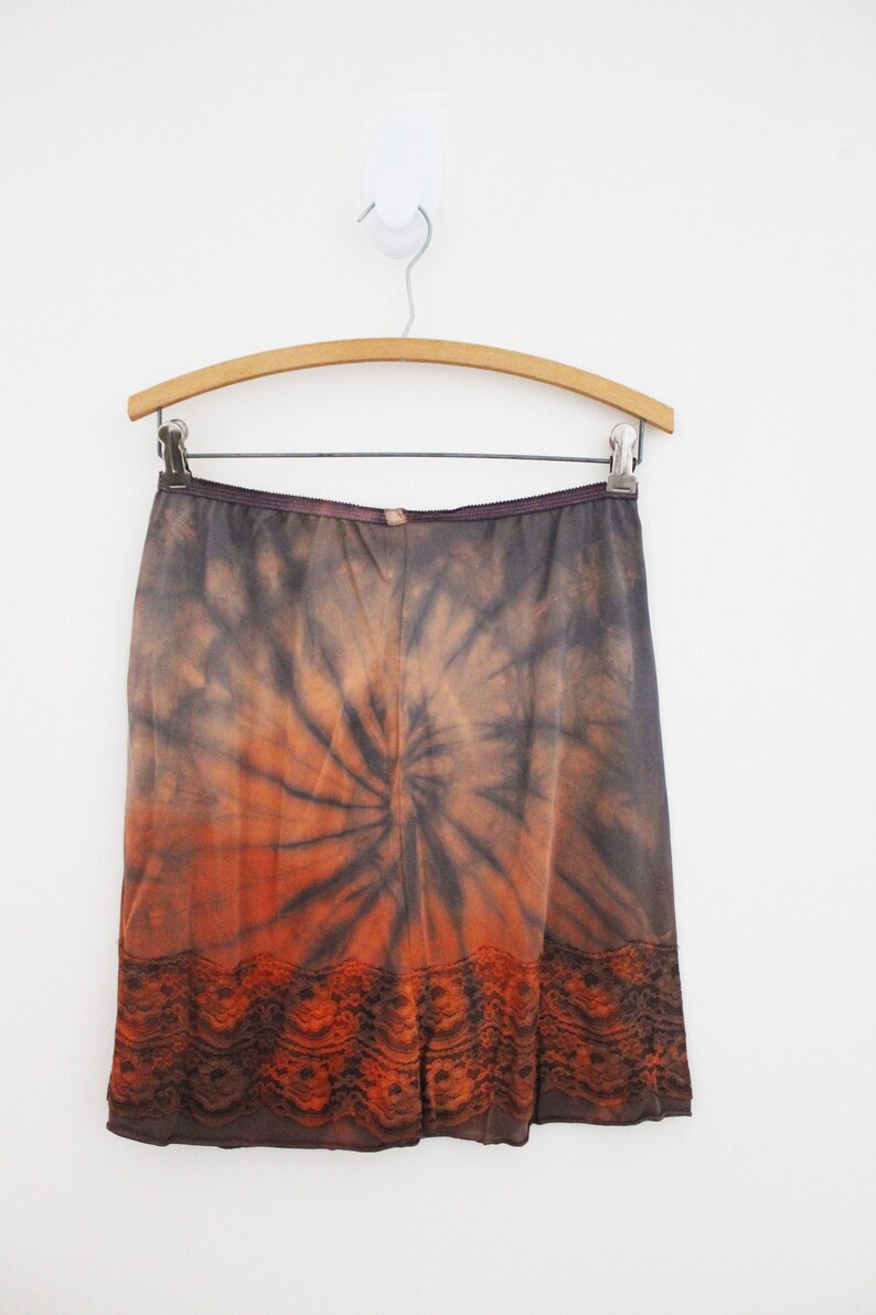 May include: A tie-dye skirt in shades of orange, brown, and gray, hanging on a wooden hanger. The skirt features a spiral pattern and a lace trim at the hem. The garment is displayed against a white background.
