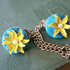 Puede incluir: Un par de broches de flores azules y amarillas conectadas por una cadena dorada. Cada broche presenta una flor amarilla con un centro de perla blanca.