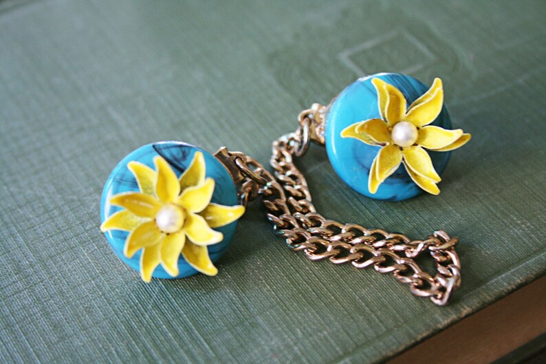 Puede incluir: Un par de broches de flores azules y amarillas conectadas por una cadena dorada. Cada broche presenta una flor amarilla con un centro de perla blanca.