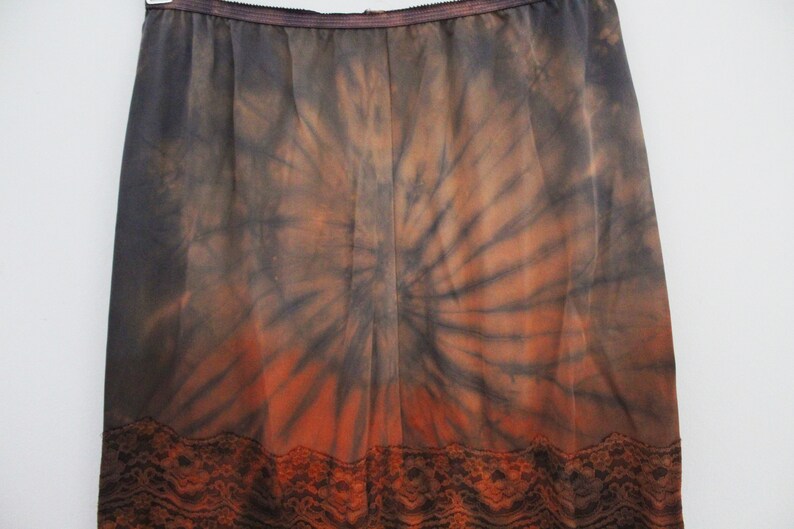 May include: A tie-dye skirt in shades of black, orange, and brown. The skirt features a spiral pattern and a lace trim at the hem. The elastic waistband is visible at the top.