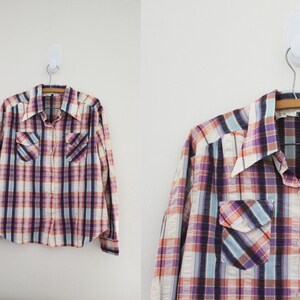 May include: A long-sleeved plaid shirt with a button-down collar. The shirt has a white, pink, purple, and blue plaid pattern. The shirt has two chest pockets.
