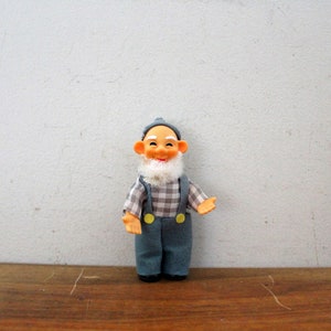 May include: A vintage doll of an elderly man with a smiling expression, wearing a blue cap, a plaid shirt, blue overalls with yellow buttons, and a white beard. The doll is standing on a wooden surface.
