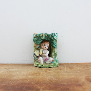 May include: A vintage ceramic vase featuring a cherubic figure seated beneath a tree. The vase is painted with green, brown, and pastel colors, and the cherub holds a butterfly. A decorative piece.