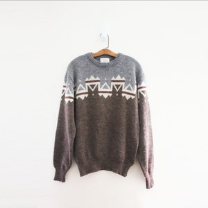 May include: A gray and brown sweater with a geometric pattern of white and maroon triangles across the chest. The sweater is hanging on a wooden hanger against a white background. The sleeves and lower portion of the sweater are brown.