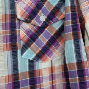 May include: A close-up of a plaid shirt with a pocket on the left side. The shirt is made of a light-weight fabric and has a relaxed fit. The colors of the plaid are blue, pink, orange, and white.