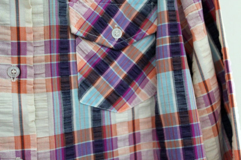 May include: A close-up of a plaid shirt with a pocket on the left side. The shirt is made of a light-weight fabric and has a relaxed fit. The colors of the plaid are blue, pink, orange, and white.