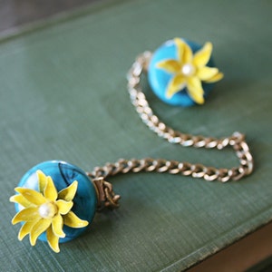 Puede incluir: Un collar de cadena dorada con dos colgantes de flores azules y amarillas. Las flores tienen un centro de perla blanca.