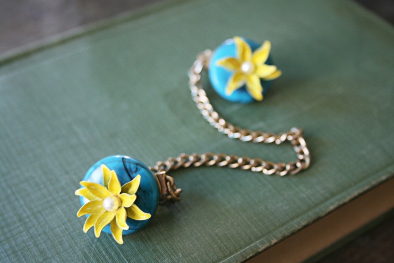 Puede incluir: Un collar de cadena dorada con dos colgantes de flores azules y amarillas. Las flores tienen un centro de perla blanca.