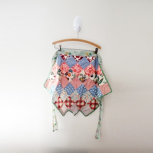 May include: A colorful, patchwork apron with a variety of patterns, including floral, diamond, and plaid designs. The apron is trimmed with green rickrack and has long ties. It hangs on a wooden hanger against a white wall.