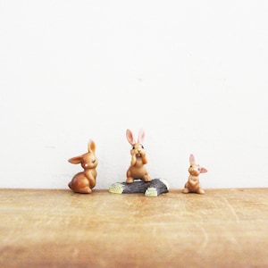 May include: Three brown rabbit figurines, one sitting on a small log, the other two standing on a wooden surface.