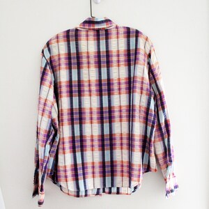 May include: A plaid button-down shirt with a white, blue, purple, and orange plaid pattern. The shirt is hanging on a white hanger against a white wall.
