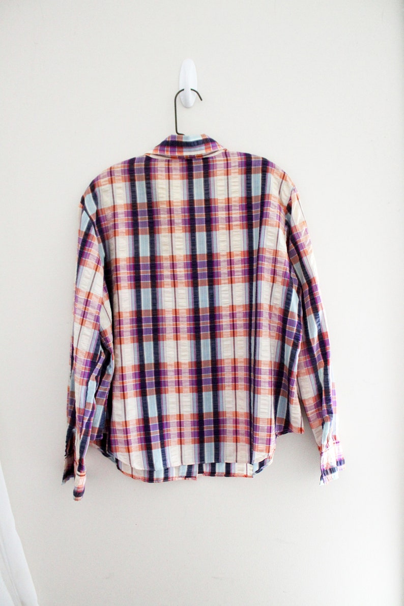 May include: A plaid button-down shirt with a white, blue, purple, and orange plaid pattern. The shirt is hanging on a white hanger against a white wall.