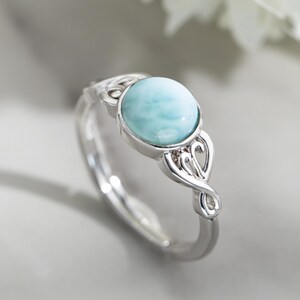 Könnte beinhalten: Ein silberner Ring mit einem hellblauen, ovalen Edelstein. Der Ring hat ein kompliziertes, wirbelndes Design am Band und um die Steinfassung. Der Edelstein hat ein marmoriertes Aussehen. Der Ring ist vor einem weichen, neutralen Hintergrund.
